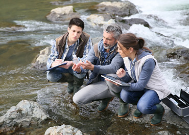 Drei Personen am Fluss schauen sich eine Wasserprobe an